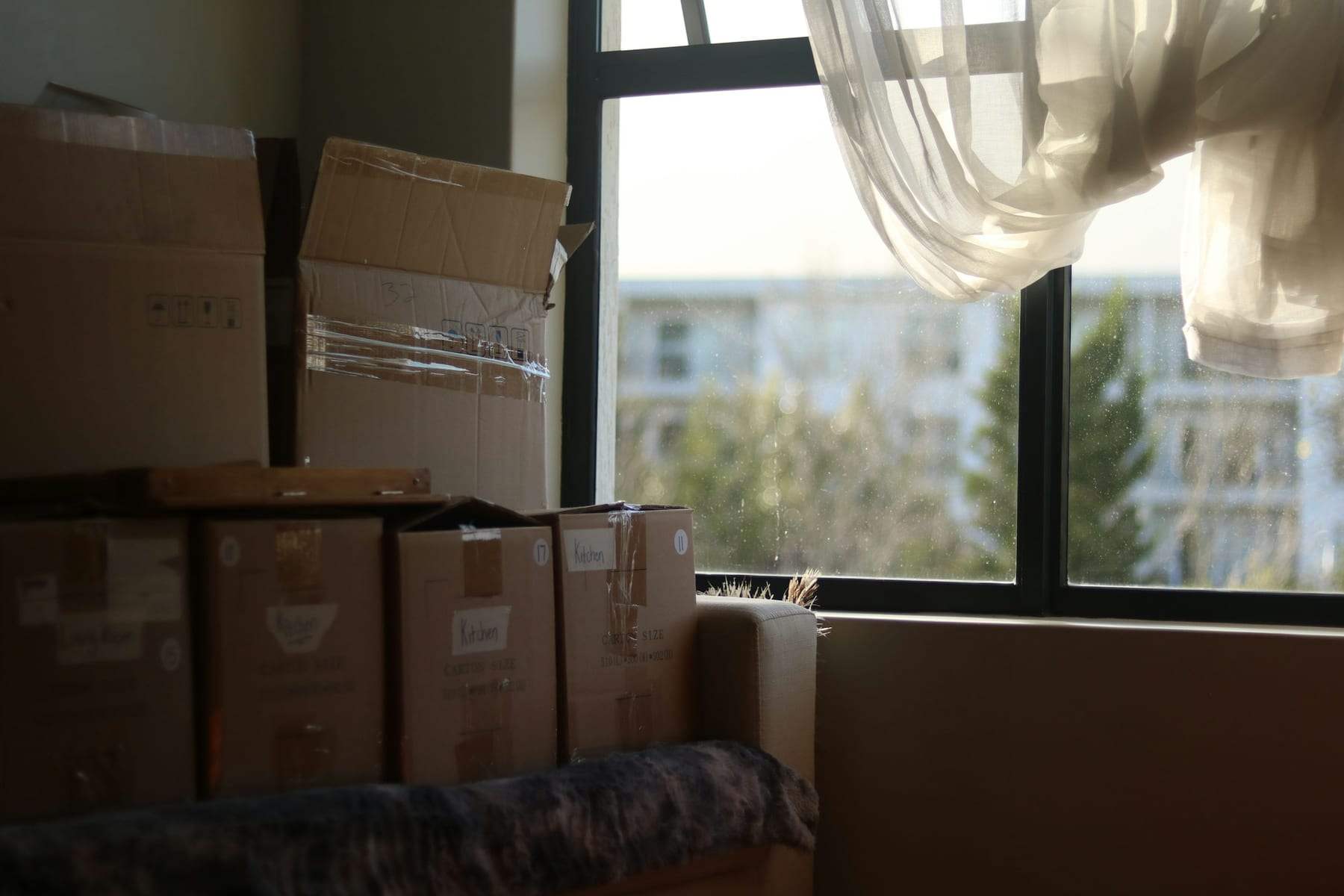 Packed cardboard boxes stacked by a sunny window with a sheer curtain