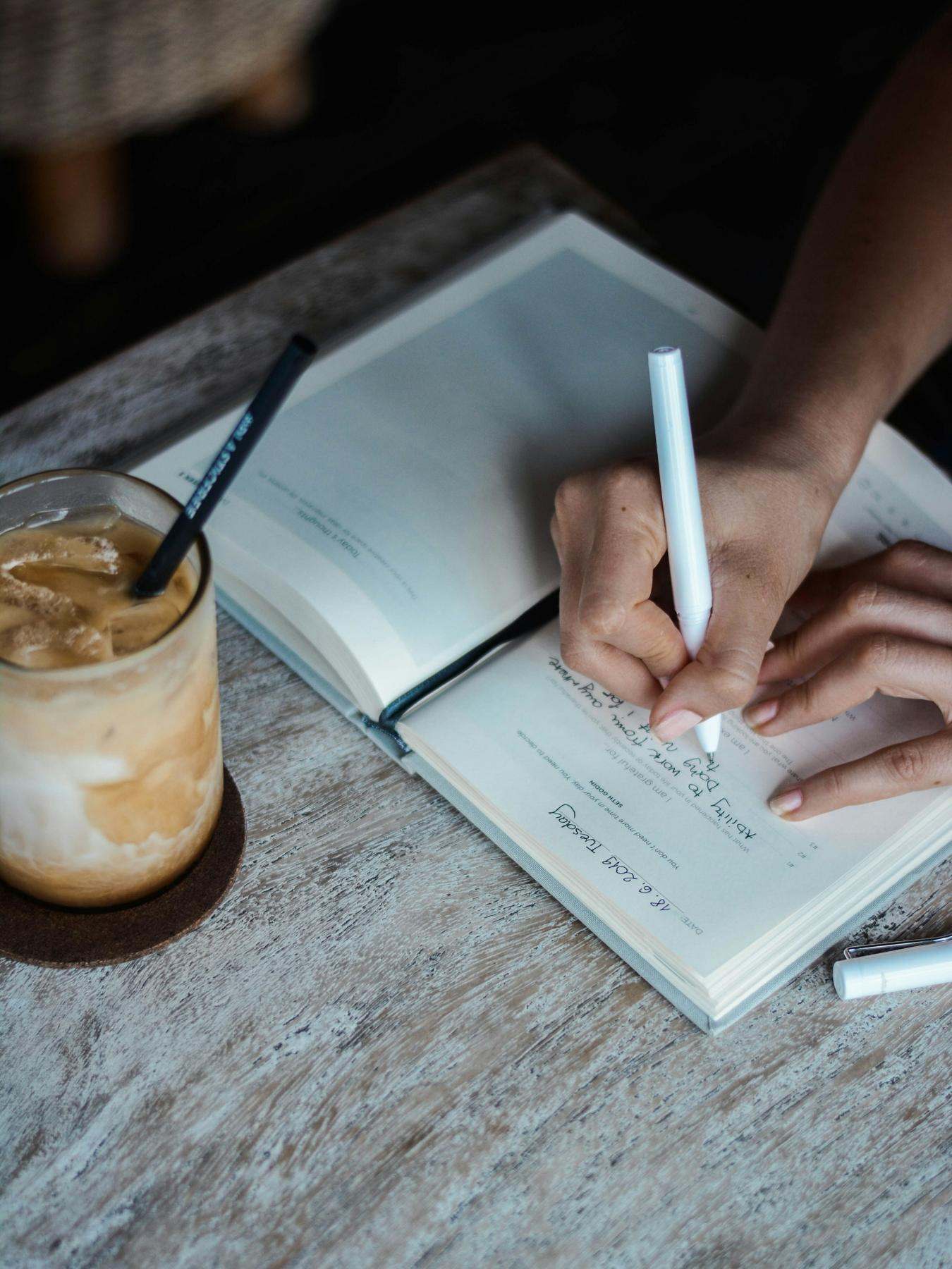 Hands writing in a notebook beside an iced coffee