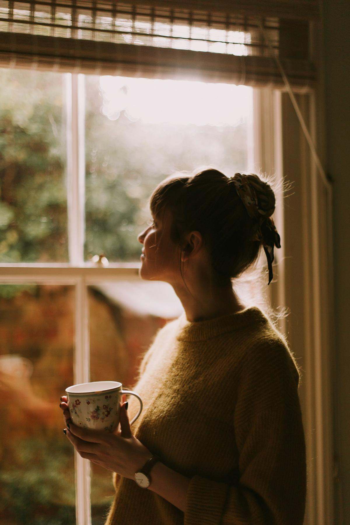 A woman holding a floral mug and looking out of a sunny window