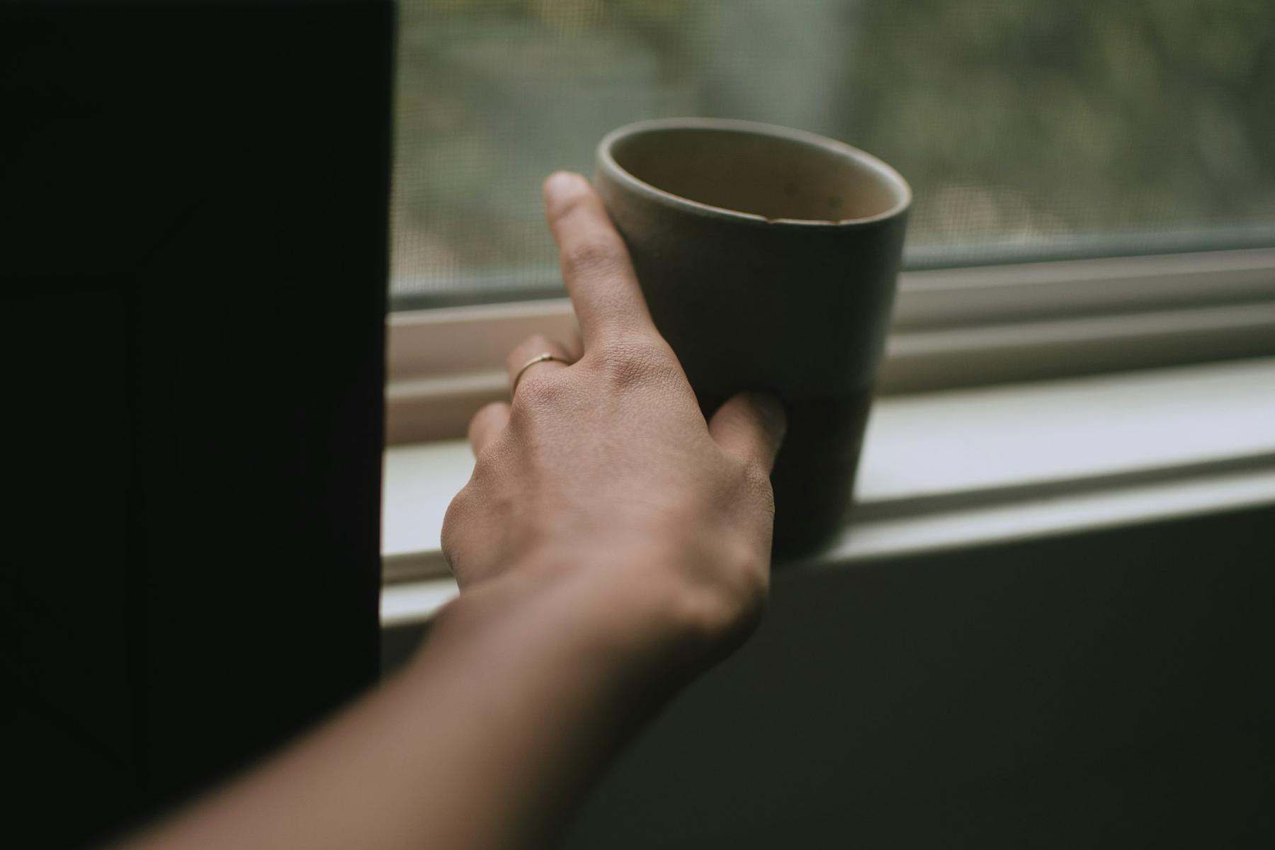 A hand resting on a mug on a windowsill in soft daylight