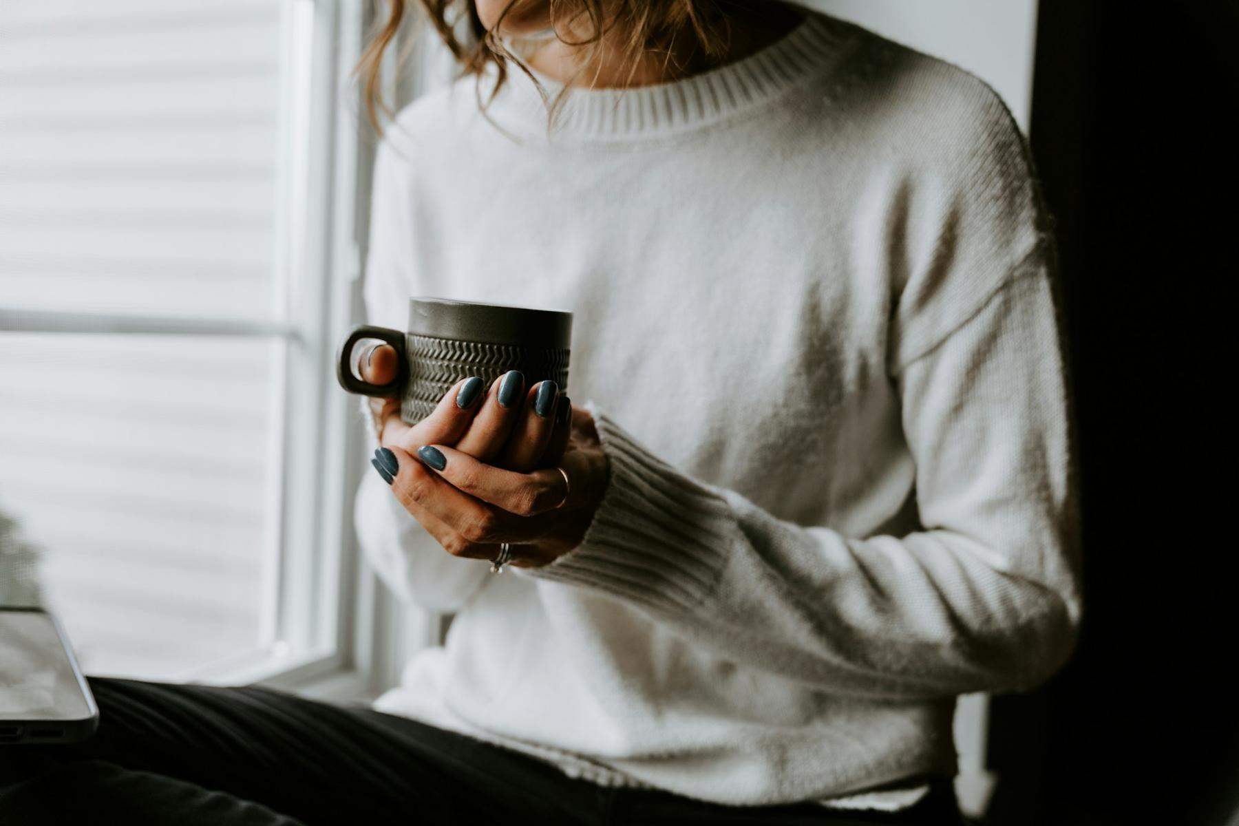 Hands holding a dark mug, wearing a cream jumper beside a window