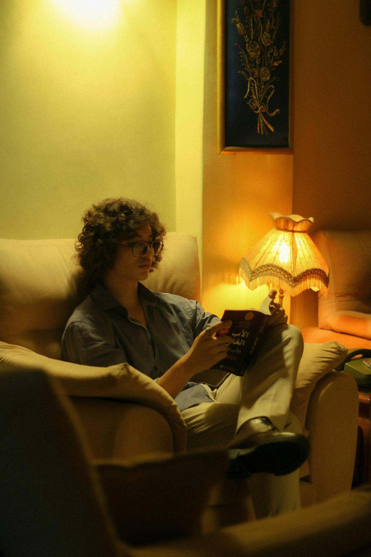 A person reading a book in an armchair under a warm lamp