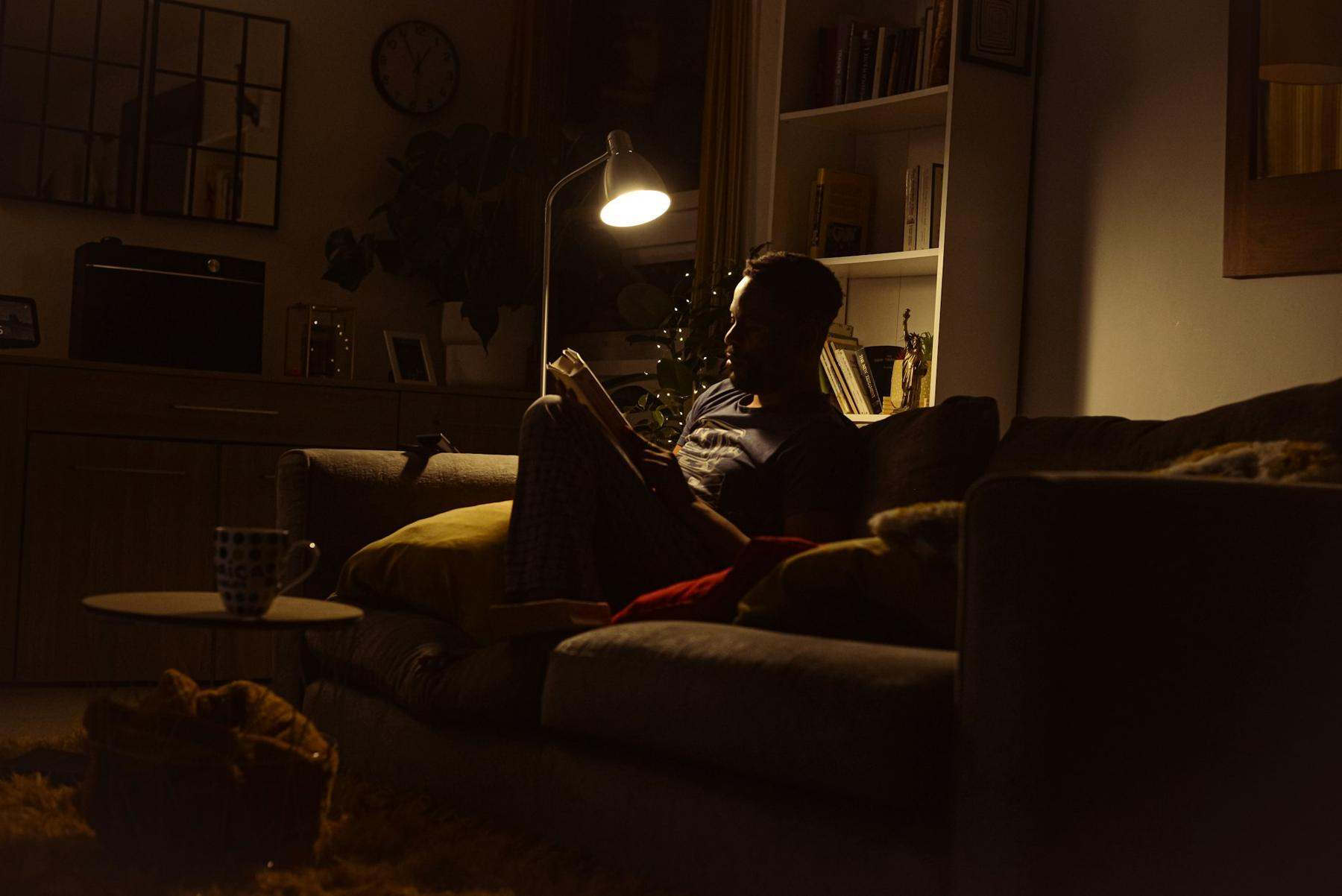 A person reading on a sofa under a lamp in a dim room