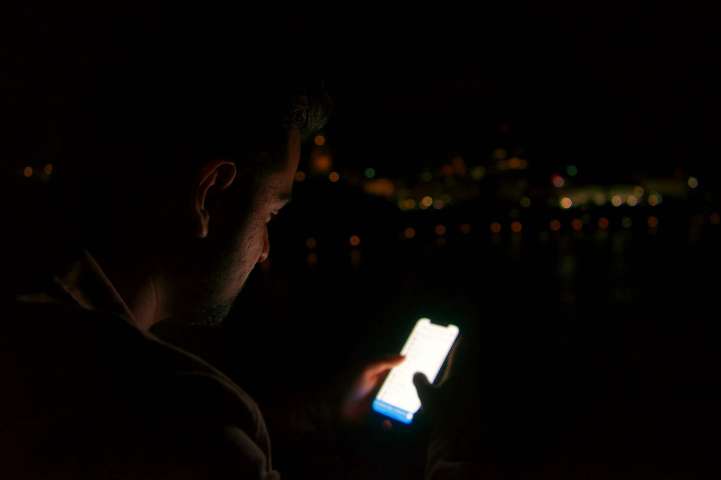 A person seen from behind looking at a phone at night, city lights beyond