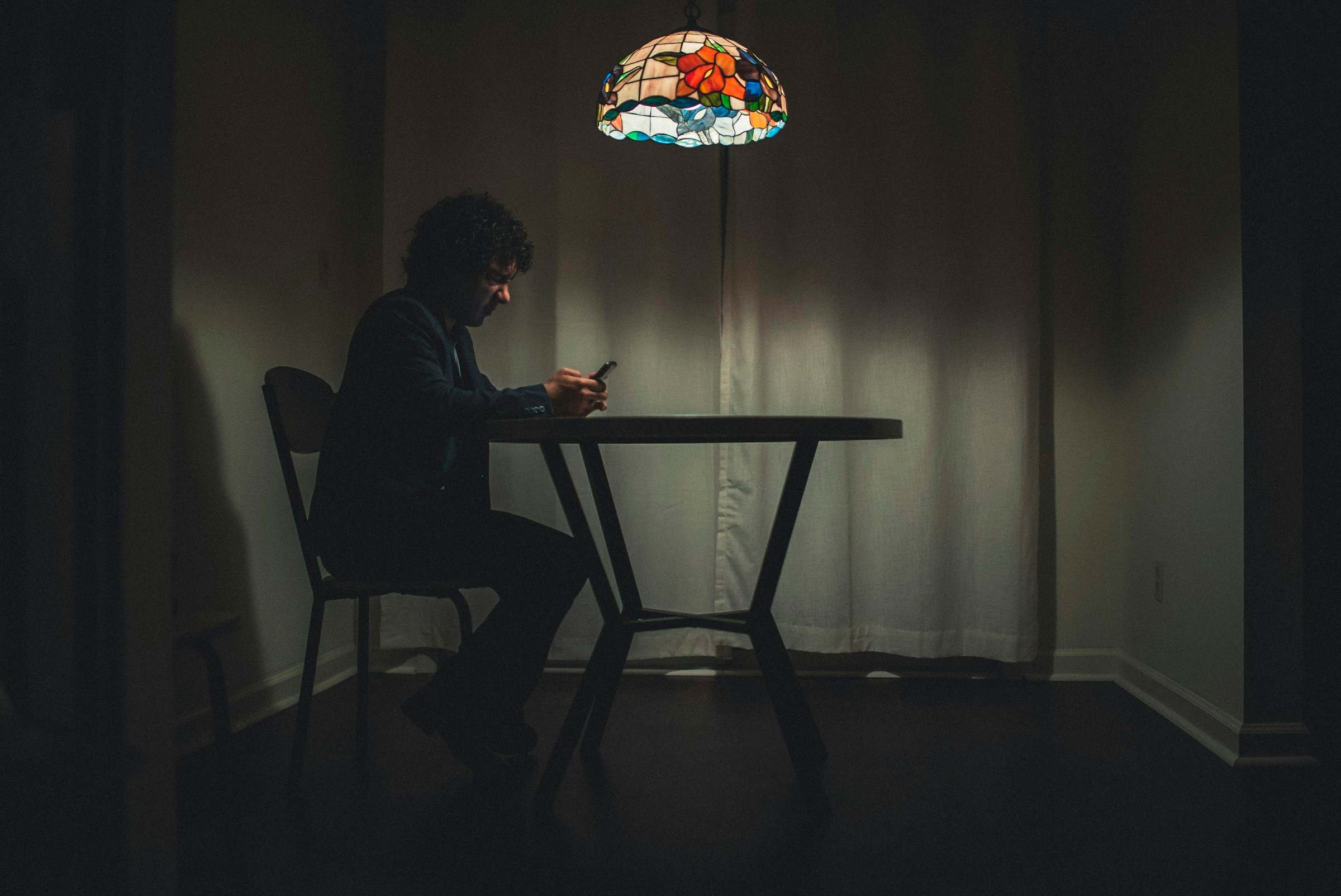 A person sitting alone at a table under a lamp, looking at a phone