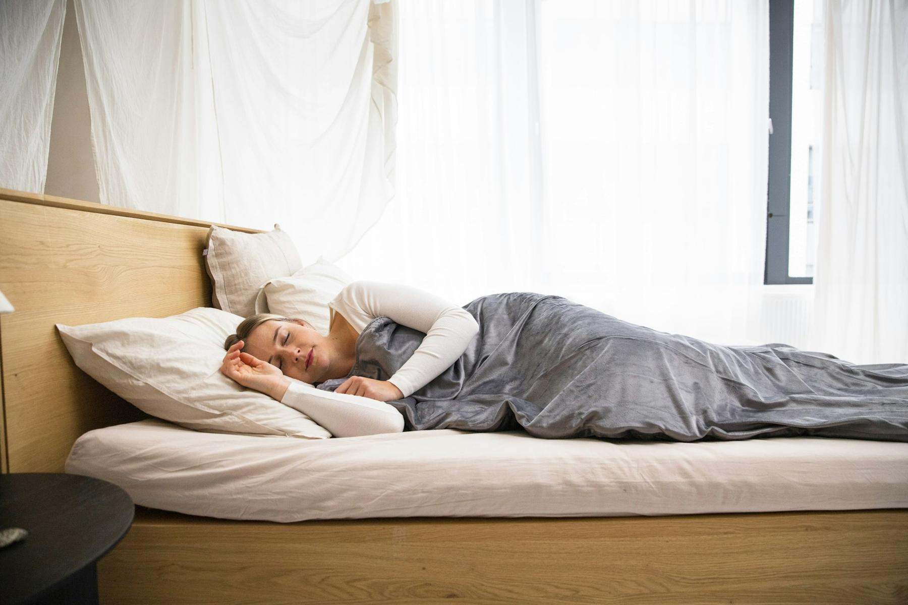 A woman asleep and smiling under a grey blanket in a bright bedroom