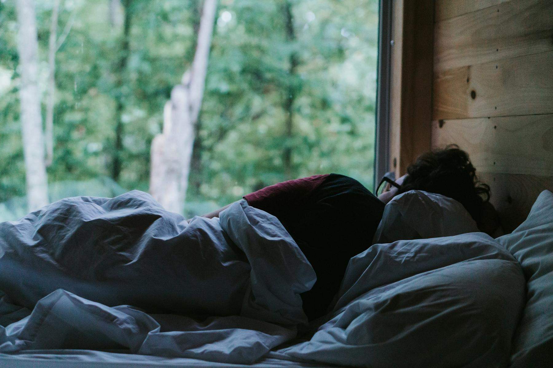 A person curled up under grey covers beside a window looking out on trees